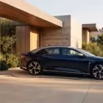 A dark blue electric sedan charges at a wall-mounted station outside a modern wood-paneled home.