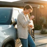 Smiling man holding car keys while leaning on a silver electric truck at sunset.