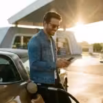 A person smiling while refueling their car at a gas station during a golden sunset, looking at their phone.