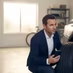 Man in a suit inspecting a car tire while holding a smartphone in a modern garage.