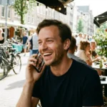A man laughs while having a video call on his phone at an outdoor cafe.