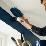 A man in white overalls carefully paints interior crown molding with dark blue paint.