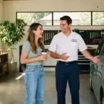 Mechanic gestures toward a hybrid car while talking to a happy customer in a garage.
