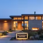 Modern luxury home exterior at dusk with large windows and a professional security sign.