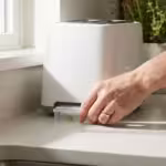 Hand removing a crumb tray from a white toaster on a bright kitchen counter.