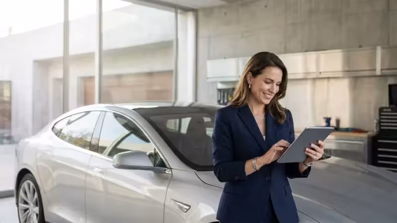Smiling businesswoman using a digital tablet next to a silver electric car in a modern showroom