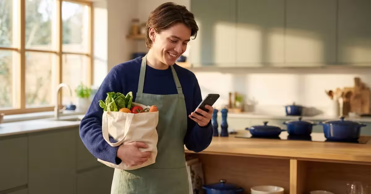 Smiling person in an apron holding fresh vegetables and using a smartphone in a kitchen.