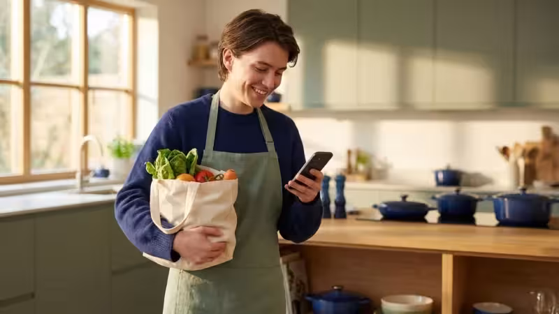 Smiling person in an apron holding fresh vegetables and using a smartphone in a kitchen.