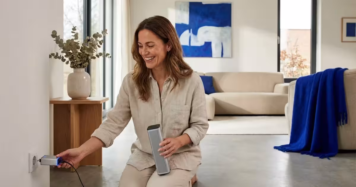Smiling woman in a modern living room plugging a portable speaker into a wall outlet.