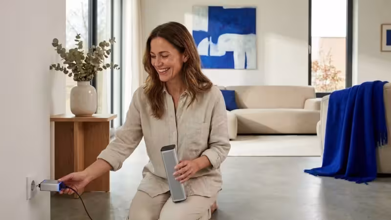 Smiling woman in a modern living room plugging a portable speaker into a wall outlet.