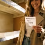 Smiling woman holding a raincheck slip for avocados in front of empty grocery store shelves.