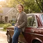 A smiling woman in a blazer leans against a vintage maroon car in a suburban driveway.