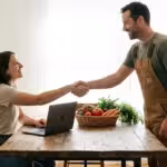 Smiling woman at a laptop shaking hands with a man in overalls over fresh vegetables.
