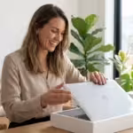 Smiling woman unboxing a new silver Apple MacBook laptop in a bright room with plants.