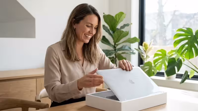 Smiling woman unboxing a new silver Apple MacBook laptop in a bright room with plants.