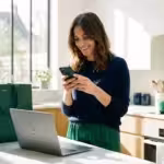 Smiling woman using a smartphone while standing at a kitchen island with an open laptop