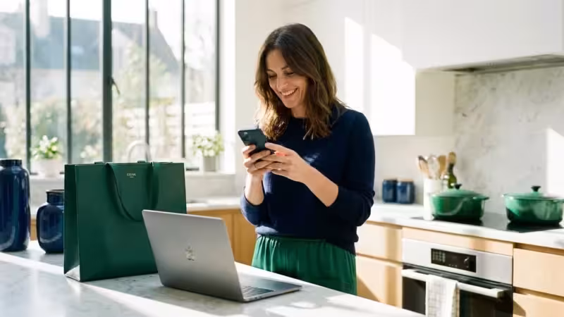 Smiling woman using a smartphone while standing at a kitchen island with an open laptop
