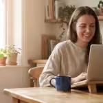 Smiling woman typing on a laptop at a wooden table in a sunlit kitchen setting.