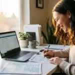 Smiling woman writing on paper documents at a bright home office desk with laptop