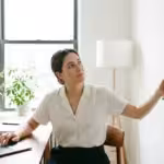 A woman sitting at a wooden desk using a laptop while measuring a white wall