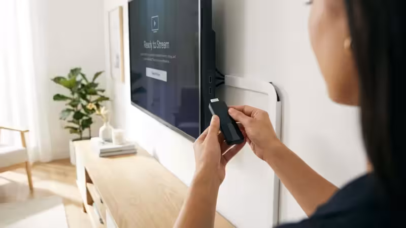 Woman inserting a black media streaming device into the side of a wall-mounted smart television.